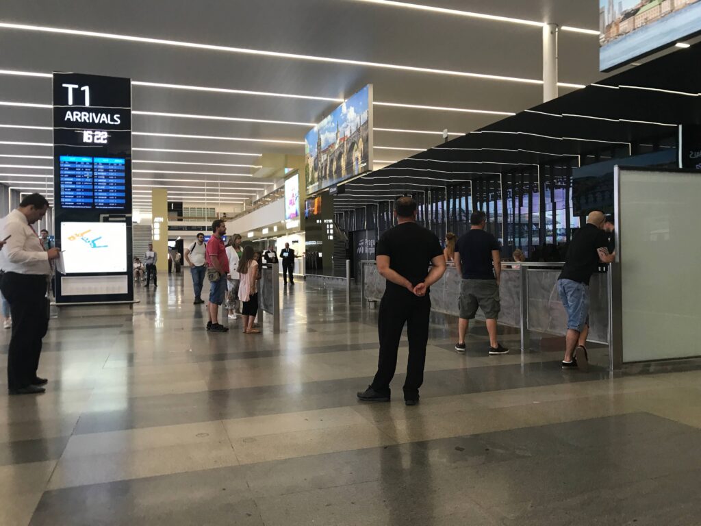 Passengers inside Prague Airport Terminal 1 arrivals hall checking flight information screens