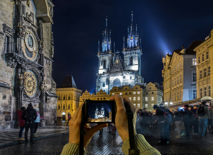 Visitor photographing Old Town Square Prague at night with the Astronomical Clock and Church of Our Lady before Týn in the background