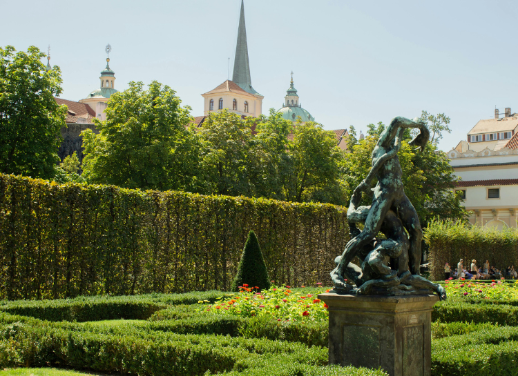 Statue surrounded by manicured hedges and flower gardens in the South Gardens of Prague Castle in Prague, Czech Republic, with historic church towers rising above the greenery in the background.
