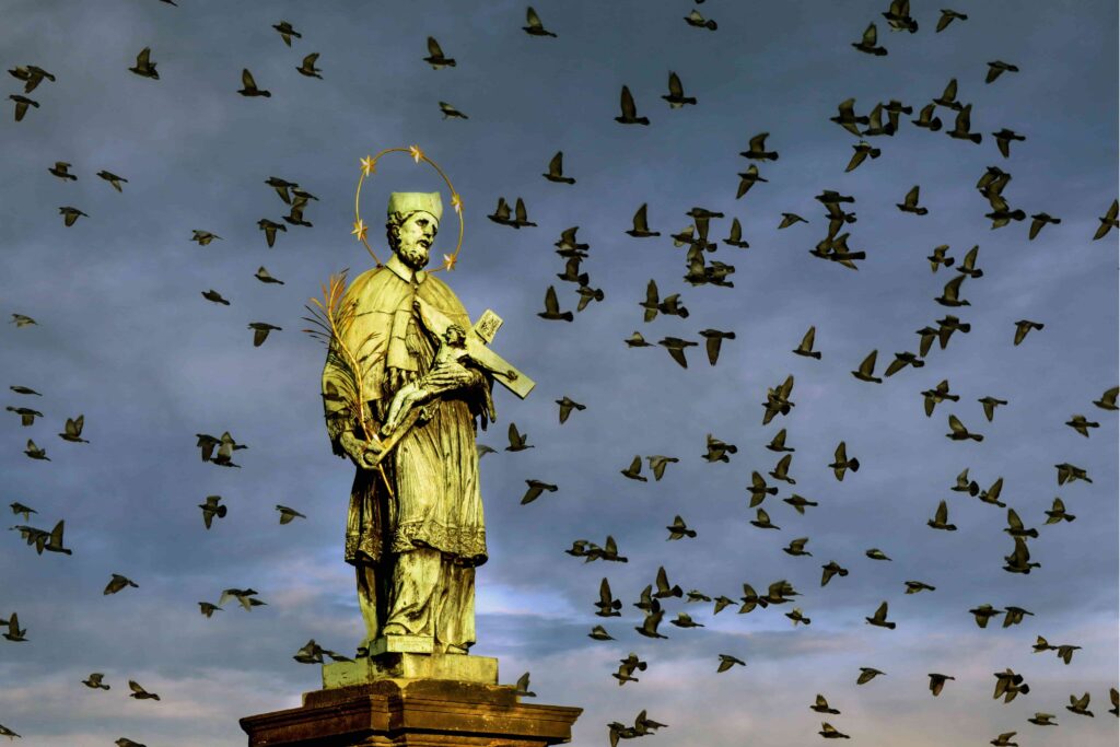 St. John of Nepomuk statue on Charles Bridge Prague, crowned with five stars and surrounded by birds, representing the legendary martyr behind one of the enduring dark stories in Prague