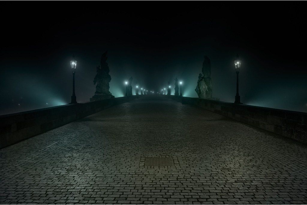 Prague Night Activities on Charles Bridge at night, with illuminated statues and historic street lamps in the fog
